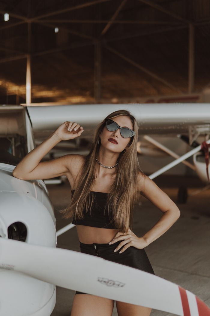 Stylish woman posing confidently by a plane in a hangar, showcasing a trendy fashion look.