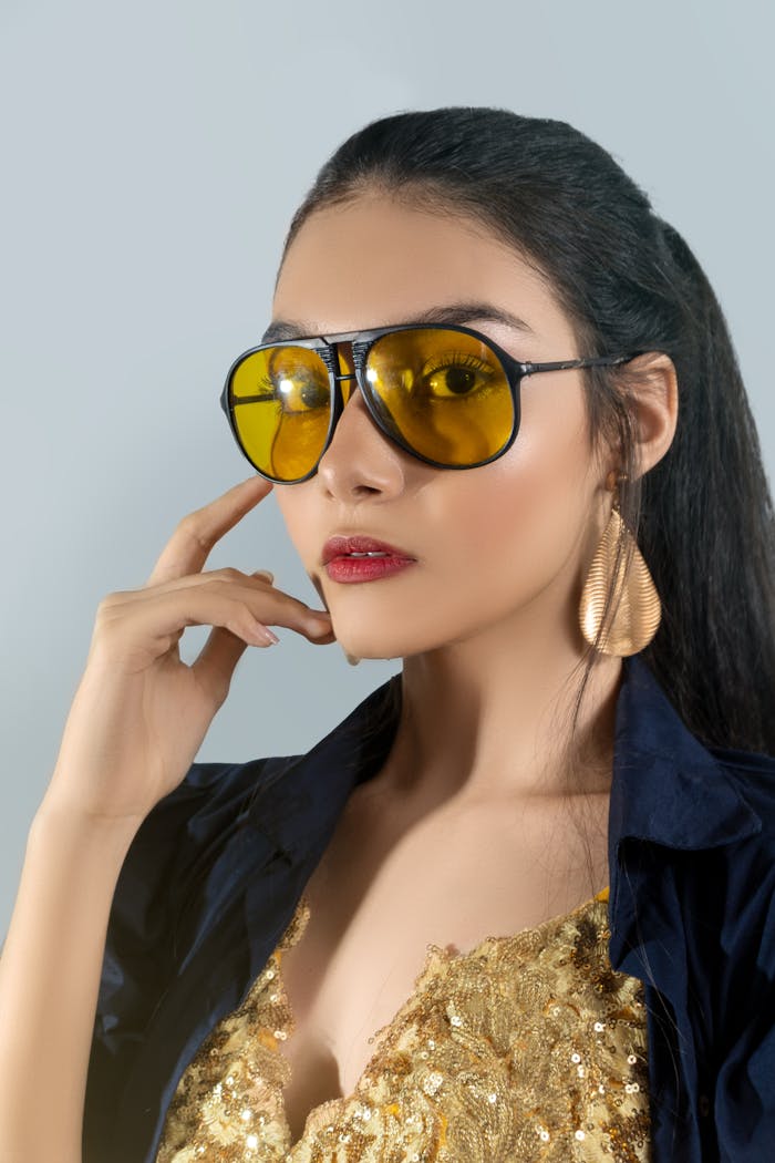 Home A fashionable young woman wearing yellow sunglasses and striking a pose in a studio setting.